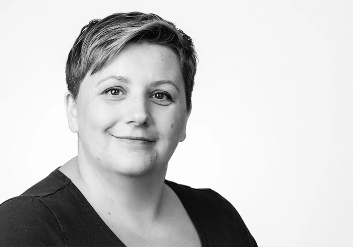 Sie hat kurze, helle Haare und schaut freundlich in die Kamera. Sie trägt ein dunkles Oberteil.