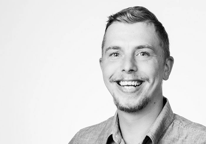 Er hat glatte, kurze Haare, einen Kinnbart und lacht freundlich in die Kamera.