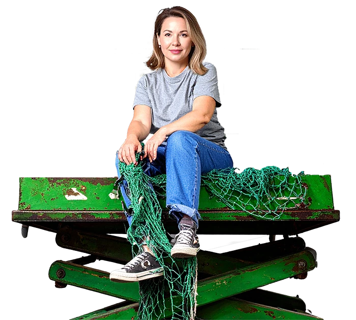 Frau in Jeans und T-Shirt sitzt auf grüner Hebebühne mit grünem Netz und blickt ruhig in die Kamera.