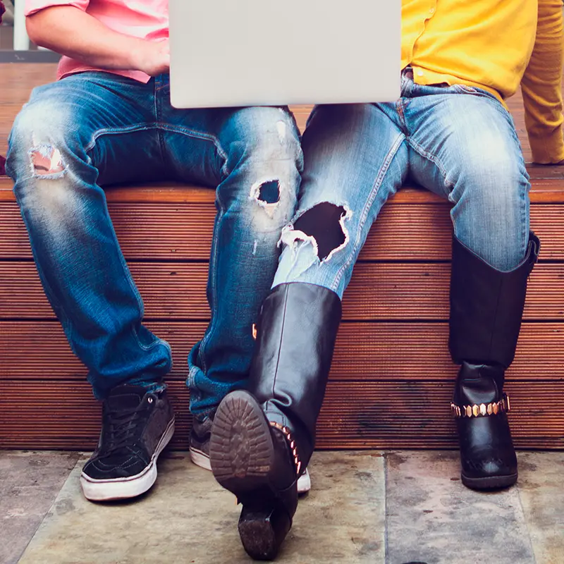 Zwei Personen in Jeans mit Laptop sitzen nebeneinander auf einer Holzbank.